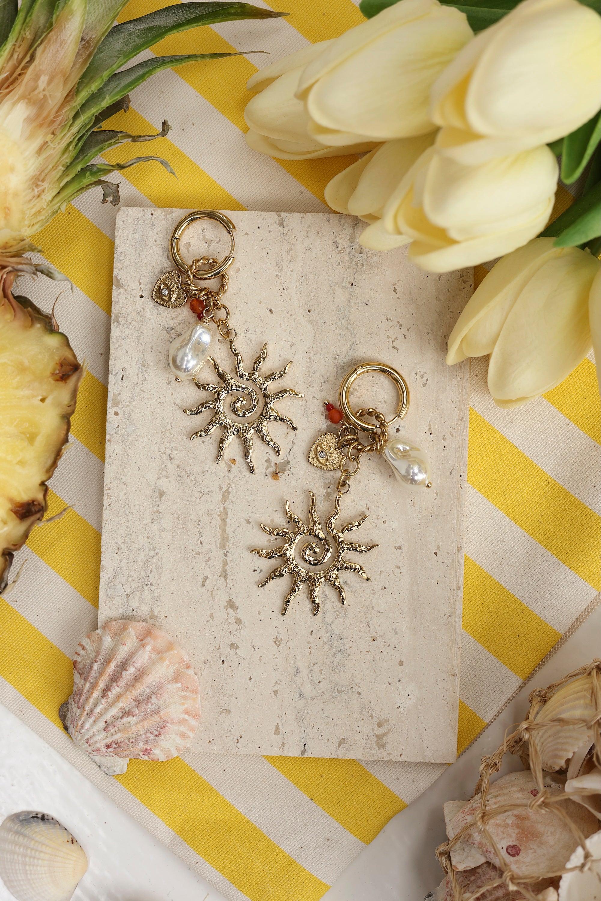 Boucles d'oreilles mini créoles dorées Ulysse avec pendentifs soleil, perle en résine et cœur orné d'un œil et d'un zircon - Bijou photographié dans un décor estival
