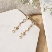 Boucles d'oreilles pendantes Célesta étoiles, lune et soleil - Bijou photographié sur un présentoir en marbre blanc à côté d'un tissu en lin beige et de fleurs blanches