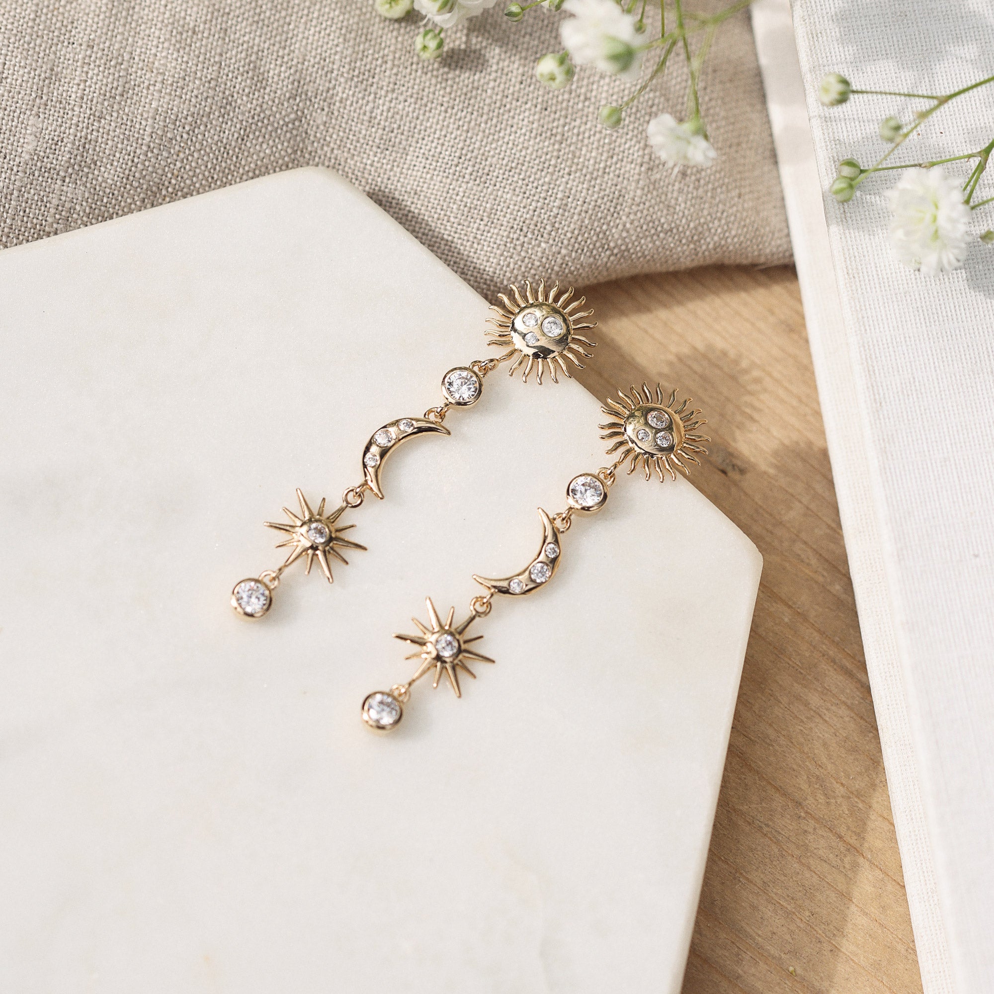 Boucles d'oreilles pendantes Célesta étoiles, lune et soleil - Bijou photographié sur un présentoir en marbre blanc à côté d'un tissu en lin beige et de fleurs blanches