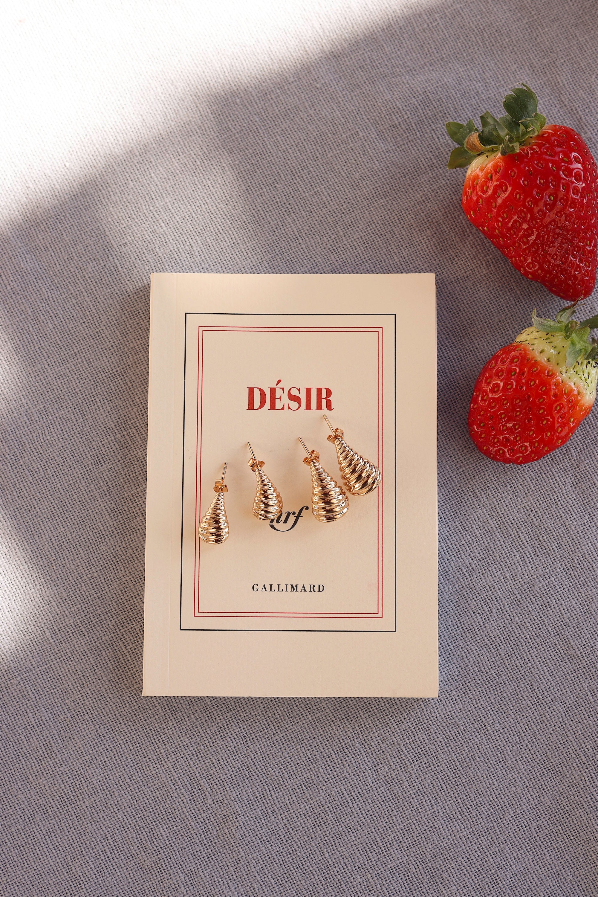Boucles d'oreilles dorées Clelia M - Bijou photographié sur livre et tissu bleu accompagné de fraise