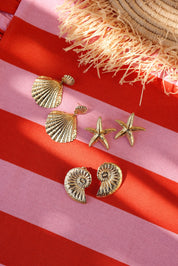 Boucles d'oreilles pendantes dorées Elyos coquillage - Bijou photographié sur tissu ligné rose et rouge avec chapeau de paille et autres boucles d'oreilles de type coquillage et étoile de mer