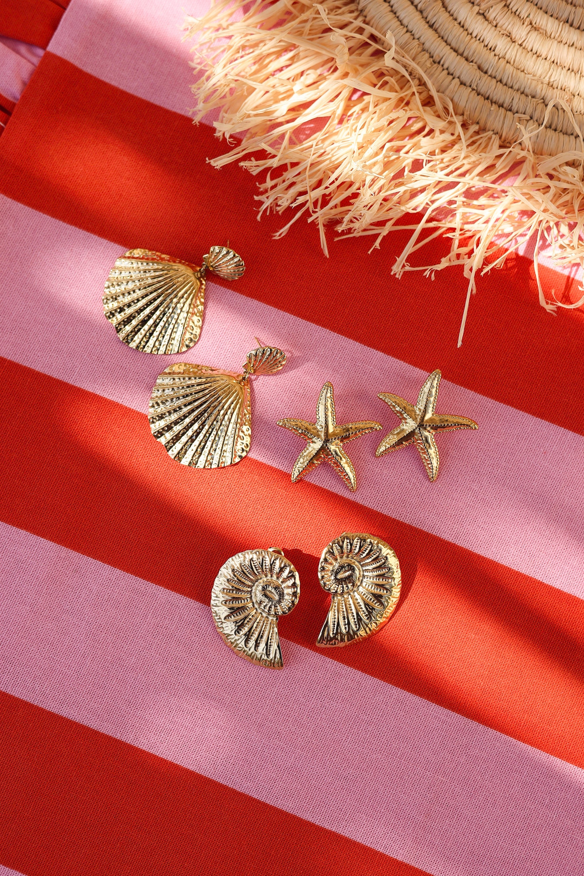 Boucles d'oreilles pendantes dorées Elyos coquillage - Bijou photographié sur tissu ligné rose et rouge avec chapeau de paille et autres boucles d'oreilles de type coquillage et étoile de mer