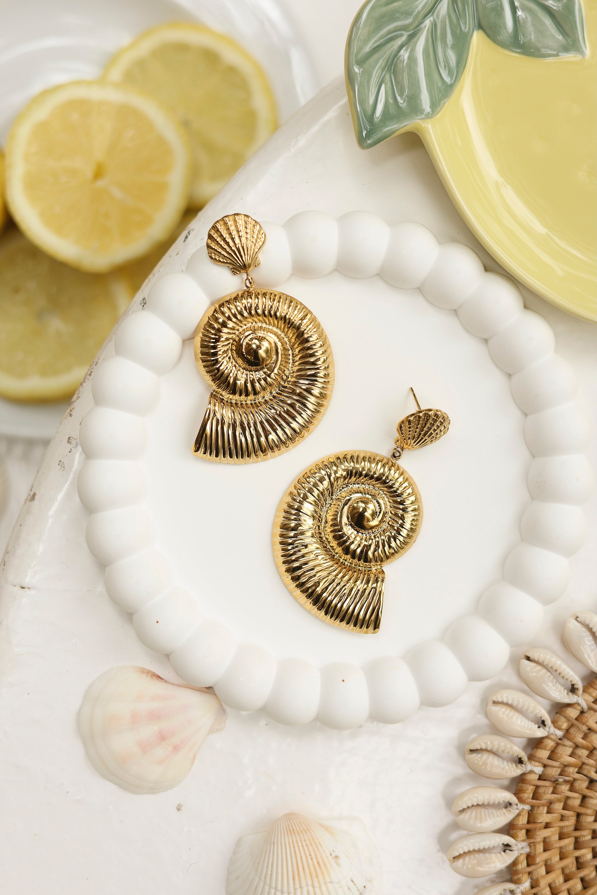 Boucles d'oreilles pendantes dorées Inaya ornées de coquillages - Bijou photographié dans un décor estival