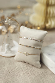 Bracelet doré Athina avec motif croix ornée de zircons - Bijou photographié sur un coussin en velours blanc cassé avec des décorations de Noël en fond