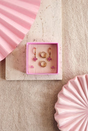 Coffret de parure de bijoux Sonia rose comprenant deux paires de mini créoles dorées ornées de zircons et une paire de puces assorties - Bijoux photographiés dans un écrin à bijou rose sur un support en marbre posé sur du lin avec des décoration rose
