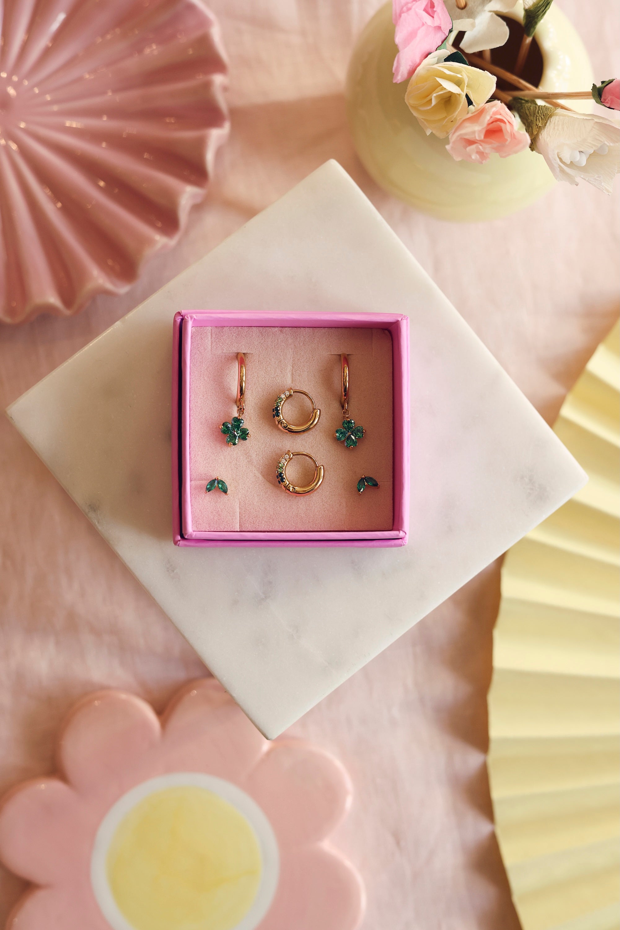 Coffret de parure de bijoux Sonia vert comprenant deux paires de mini créoles dorées ornées de zircons et une paire de puces assorties - Bijoux photographiés sur un bloc en marbre blanc avec des décorations roses et jaunes