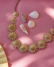 Collier doré Darina avec motifs floraux en relief - Bijou photographié sur un tissu rose avec des coquillages