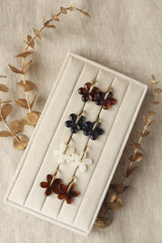 Créoles dorées Shauna beige avec pendentif fleur en acétate - Bijou photographié sur un présentoir blanc crème posé sur un tissu en lin avec des feuilles décoratives dorées