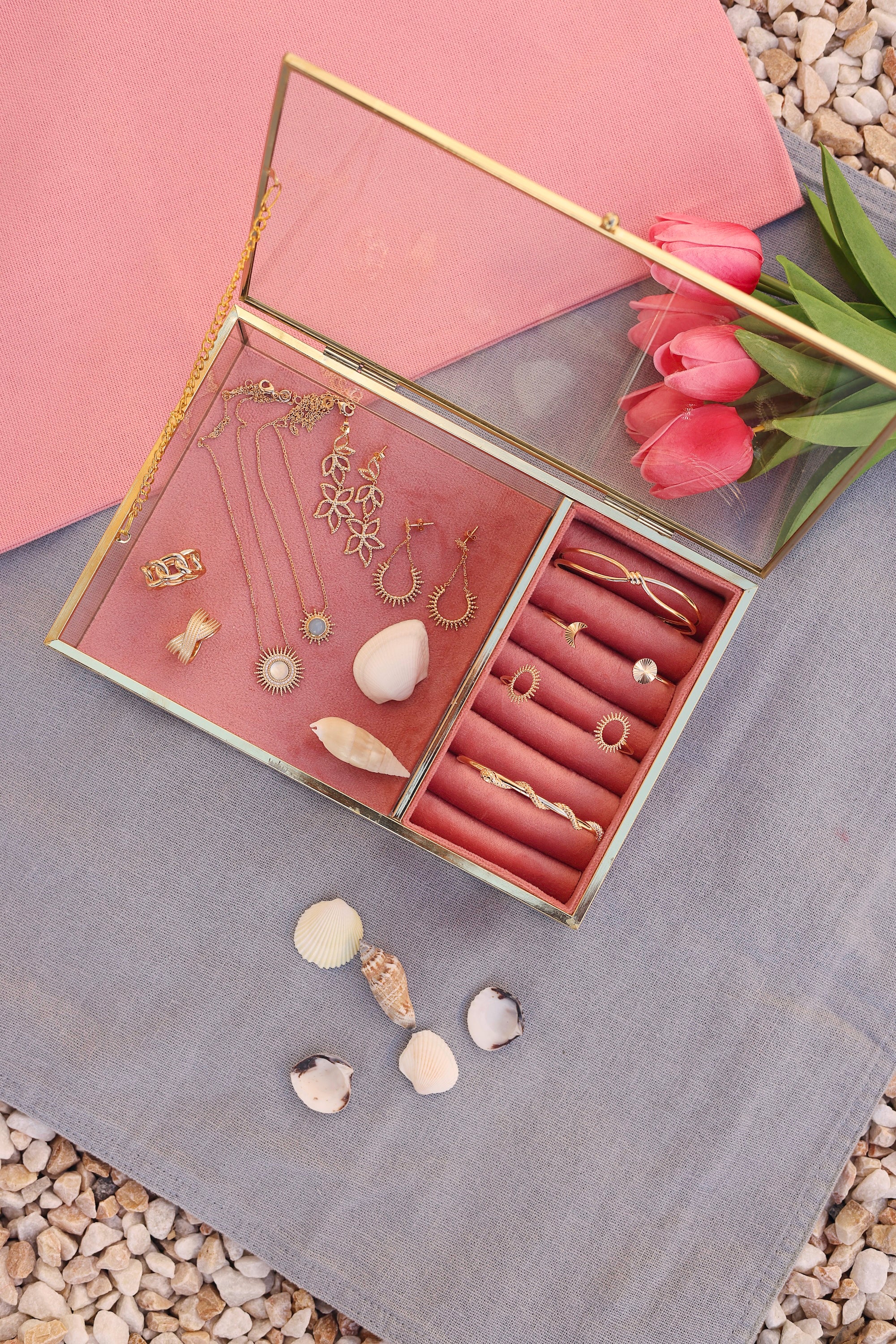 Jonc doré Kiane nœud - Bijou photographié dans une boite à bijou en velours rose et en verre aux bords doré posé sur un tissu bleu posé sur des cailloux avec des tulipes et des coquillages